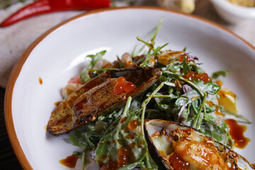On a dark countertop, a light linen napkin, on a cutting board  Salad with mussels, rucola, red caviar, shrimps in a round dish, cutlery, pepper-salt steam, whole red pepper, hot pepper pod