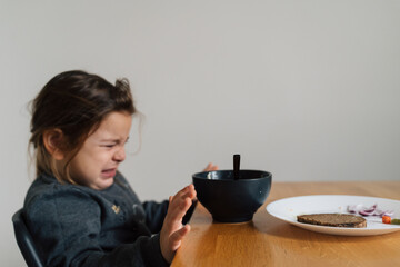 Unhappy child girl eats soup from black bowl with bread and onion. Lifestyle photo of kid in kitchen having a meal, Screaming kid. Picky eater - obrazy, fototapety, plakaty