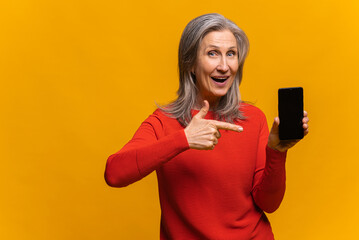 Cheerful mature gray-haired woman showing smartphone with empty blank screen for inserting advertise, point finger at display. Happy senior woman recommends holiday deal