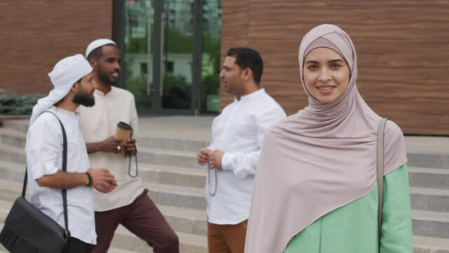 Waist-up of beautiful Caucasian Muslim woman wearing hijab, smiling and looking on camera, standing in foreground of three men talking by office building
