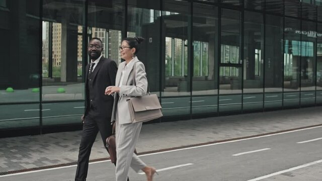 PAN shot of elegant African-American businessman with umbrella and Asian businesswoman in formal wear walking down street in financial district of city and chatting