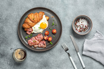 Breakfast croissant sandwich with fried egg, bacon, tomato, microgreens, coffee. Gray grunge background.Top view, flat lay