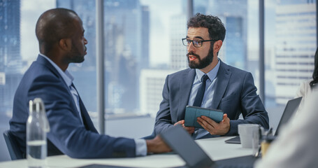 Modern Multi-Ethnic Office Conference Room Meeting: Caucasian Top Manager Talks to Black Executive, Brainstorming, Forming Strategy, Using Tablet Computer to Find Solution. Medium Portrait