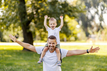 Father and son play in nature. They imagine planes while Dad carries the boy on his shoulders. They have fun and have fun in the park, the same casual clothes. Nice family moment