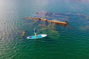Woman on paddle board, sup next to Abandoned broken shipwreck sticking out of the sea