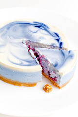 Piece of blue and white cheesecake with blueberry, cherry and black currant sauce inside on white plate on white table
