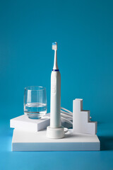 Electric toothbrush with glass of water on white podium on blue background. Modern oral hygiene...