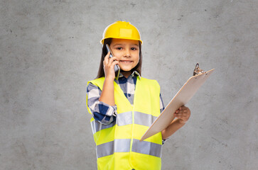 building, construction and profession concept - smiling little girl in protective helmet and safety vest with clipboard calling on smartphone over grey concrete wall background