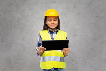 building, construction and profession concept - smiling little girl in protective helmet and safety vest with tablet pc computer over grey concrete wall background