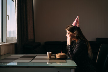 Cute thoughtful woman in party hat looking at festive cake with candle. Lonely woman freelancer...
