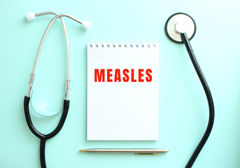 On a blue background, a stethoscope and a white notepad with the words MEASLES.