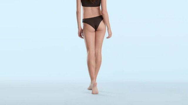 Horizontal medium shot of young slim good-looking woman in black underwear walking away from camera on tiptoes on pale blue background | Leg skin care concept