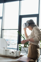 Middle aged businesswoman using vr headset near paper folder and models of buildings in office.