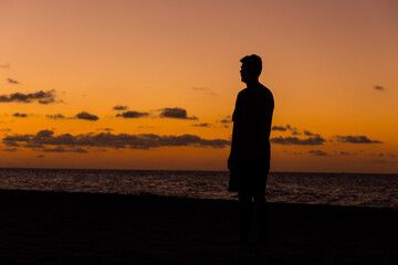 Fototapeta premium Man standing on a tropical beach at twilight silhouetted against a vivid orange ocean sunset