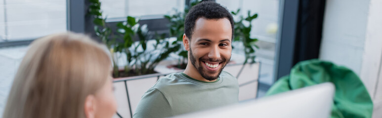 happy african american man looking at blonde colleague in office, banner.