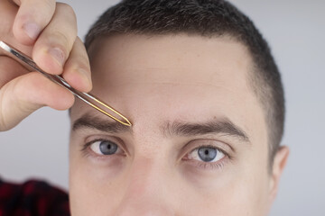 Man plucking eyebrows. Close-up of removing excess hair with tweezers. Isolated on white background. A male looks into the frame and it feels like he is looking in the mirror