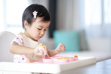 Asian cute little girl holding fork and trying to eat food sitting on the white high chair in the morning at home with copy space.