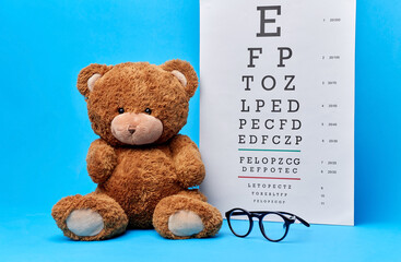 vision, health and childhood concept - brown teddy bear with glasses and eye chart over blue...