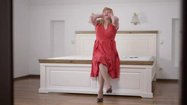 Stressed troubled Caucasian transgender woman closing ears with hands sitting on bed freaking out. Wide shot nervous LGBTQ person indoors in bedroom at home. Intolerance and self perception