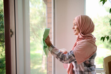 Waist-length side portrait of a cheerful happy young Arab Muslim woman with covered head in hijab washing windows while enjoying spring cleaning in house or apartment, copy space