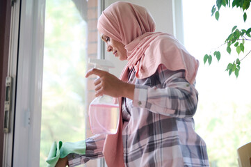 Middle aged beautiful Muslim woman with her head covered in pink hijab washing windows during...