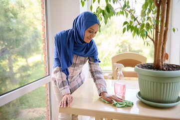 Smiling cheerful delightful beautiful Arabic Muslim woman with covered head in blue hijab wiping dust on the table in the veranda during spring cleaning. Housekeeping, chores, cleaning concept