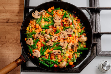 The vegetable mixture is fried in a frying pan. A mixture of carrots, green beans, peas and mushrooms is fried in a cast-iron frying pan. The process of cooking vegetarian food.