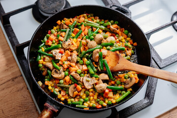 The vegetable mixture is fried in a frying pan. A mixture of carrots, green beans, peas and mushrooms is fried in a cast-iron frying pan. The process of cooking vegetarian food.