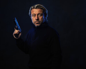 Shadowy night portrait of a confident man with a gun in his hand with blond hair, a stubble beard and a black wool sweater.