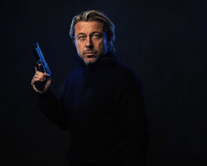 Shadowy night portrait of a confident man with a gun in his hand with blond hair, a stubble beard...