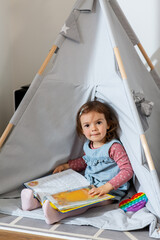 childhood, hygge and people concept - little baby girl reading book in kid's tent or teepee at home