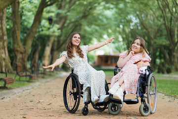 Pleasant young women with disability smiling and taking fun during summer walk at green city park. Female wheelchair users enjoying warm days on fresh air.