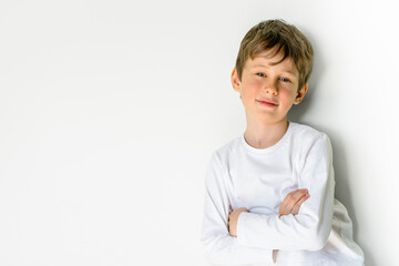 Portrait of an 8 year old boy wearing a white T-shirt with copy space. The boy with folded arms looks skeptically at the camera. Personal defense mechanisms - obrazy, fototapety, plakaty
