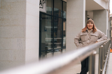 Happy smiling overweight woman in warm hat and jacket standing posing near railing of office building at city street in cloudy autumn day. Frozen obese female waiting for someone outdoors.