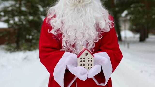 House, miniature cottage in hands of Santa Claus outdoor in snow. Deal for real estate, purchase, construction, relocation, mortgage. Cozy home, Delivery of gifts. Merry Christmas, new year booking