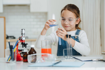 Smug little girl doing home science project, pouring blue liquid into a flask. She is wearing...