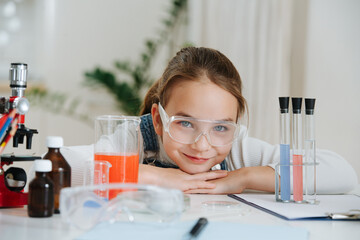 Portraitof a shcoolgirl in plastic glasses doing home science project. She is lying on the table, leaning on her hands. She has glass chemical equipment and microscope