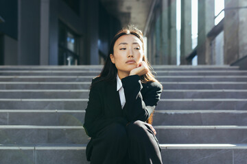 Young beautiful asian woman upset and sad sitting on the stairs of the office center, depressed...