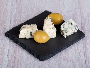 A piece of cheese with blue mold with large green olives on a slate board. Light wooden background