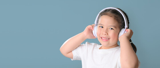 Panoramic shot of happy cute little asian girl  listening music in headphones over blue background. music, technology and people concept.