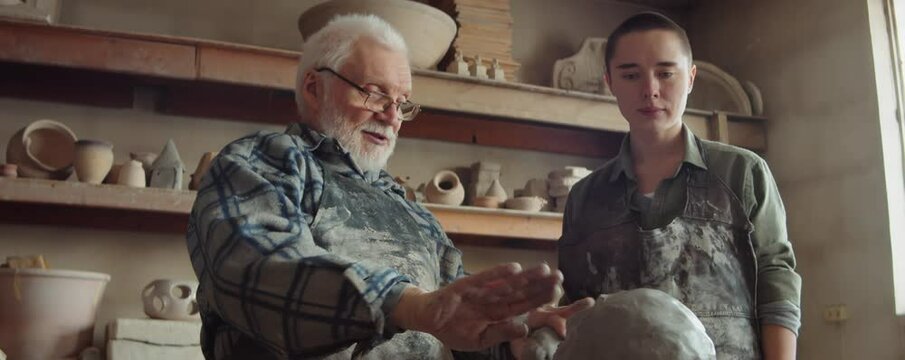 Experienced elderly sculptor teaching young female apprentice how to create clay head during lesson in pottery studio