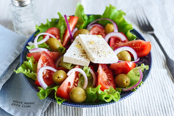 Greek salad of fresh vegetable with tomatoes, lettuce, olives, red onion and feta cheese in bowl on white striped linen tablecloth. Selective focus