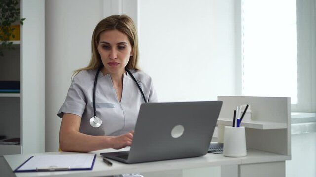 Doctor telehealth remote service. Blonde woman internist in grey uniform with spbd stethoscope writes email looking at papers by laptop in hospital