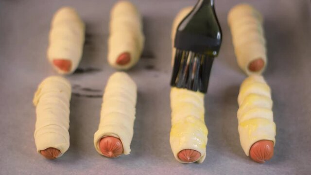 Making pigs in a blanket. Covering sausages wrapped into raw puff pastry with egg wash prepared to be baked. Homemade fast food