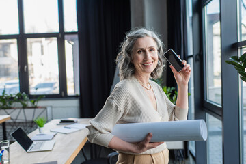 Cheerful mature businesswoman holding smartphone with blank screen and blueprint in office.