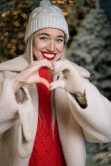 Love smiling happy face. Portrait of blonde beautiful woman in warm winter clothes hat mittens and red jumper looking at camera showing heart hand symbol. Romantic mood. Evening snowy forest magic