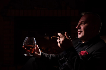 A mature man with glasses drinks brandy and smokes a cigar while sitting in an armchair by the...