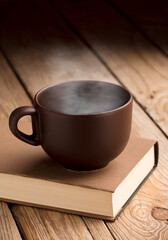 Brown cup of coffee with smoke on reading book, rustic wooden table, at sunrise. 
