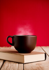 Brown coffee cup with smoke on reading book, rustic wooden table, red background.