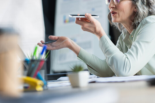 Cropped view of middle aged businesswoman recording voice message on mobile phone near papers in office.